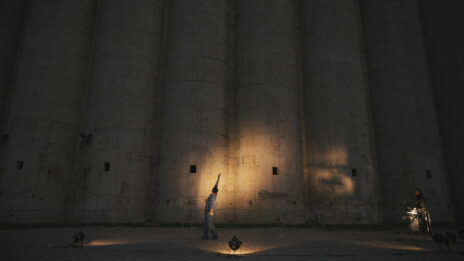 Le danseur se tient devant un très grand mur en béton, dehors. Il fait presque noir mais il y a un projecteur pointé vers lui.