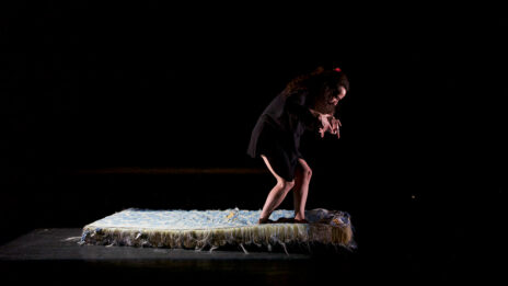 Racha Baroud debout sur un matelas au milieu de la scène, dans la pénombre.