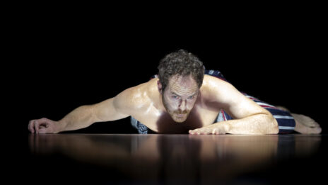 Photo de Boris Charmatz sur scène. Il rampe sur le sol dans la pénombre. On voit une partie de son reflet sur le sol. Il a l'air de se regarder dedans.