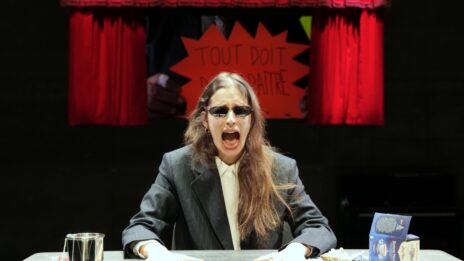 Photo de Stéphanie AFlalo sur scène, elle est assise devant un écran où passe une vidéo où est écrit "tout doit disparaître" au feutre sur une pancarte. Elle semble crier, elle a les deux mains posées à plat sur la table.